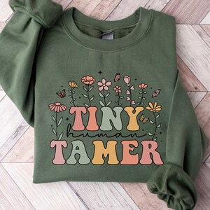 May include: A green sweatshirt with a floral design and the text "TINY human TAMER" in colorful retro lettering.