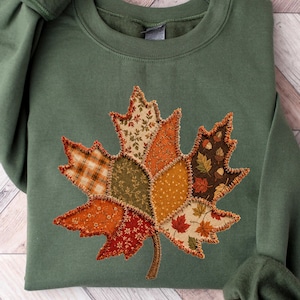 May include: A military green crewneck sweatshirt featuring a large, central maple leaf graphic composed of various patchwork-style fabric patterns in shades of orange, brown, yellow, and green, with visible stitched edges around the leaf segments.