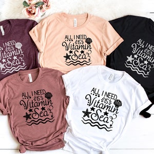 May include: Five t-shirts in different colours with the text "All I need is Vitamin Sea" in black lettering with a seashell, starfish, and wavy lines.