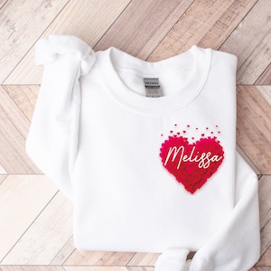 May include: White sweatshirt with a pink heart made of smaller hearts. The heart has the name "Melissa" written inside.
