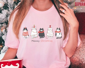 May include: A light pink t-shirt featuring a row of cartoon cats decorated for Christmas. The cats are wearing festive accessories like scarves and Christmas tree lights. The text "Meowy Christmas" is printed below the cats.
