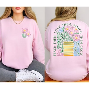 May include: A light pink sweatshirt with a floral design on the front and back. The front features a small floral graphic, while the back displays a larger illustration of flowers in a vase with the text "TEACH THEM, LOVE THEM, WATCH THEM GROW."