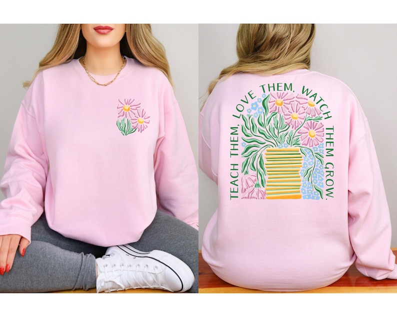 May include: A light pink sweatshirt with a floral design on the front and back. The front features a small floral graphic, while the back displays a larger illustration of flowers in a vase with the text "TEACH THEM, LOVE THEM, WATCH THEM GROW."