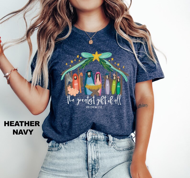May include: A navy blue t-shirt with a colorful illustration of the nativity scene. The text "The greatest gift of all" and "Matthew 1:21" are printed on the shirt.