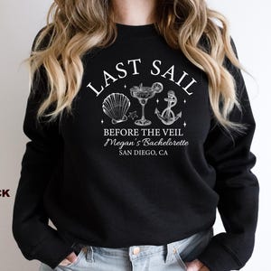 May include: Black sweatshirt with white text and nautical-themed graphics. The text reads "LAST SAIL BEFORE THE VEIL Megan's Bachelorette SAN DIEGO, CA." Graphics include a seashell, cocktail, and anchor. The word "BLACK" is printed on the left side.