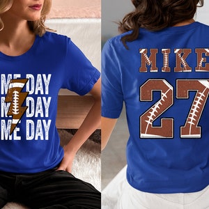 May include: Two blue t-shirts with a football theme. One shirt says "GAME DAY" three times with a lightning bolt graphic. The other shirt says "MIKE" and "27" with a football graphic.