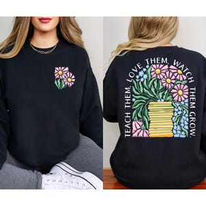 May include: Black sweatshirt with a floral design on the front and back. The front features two pink daisies with green leaves. The back has a larger floral arrangement with the text "TEACH THEM, LOVE THEM, WATCH THEM GROW."