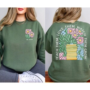 May include: Green sweatshirt with a floral design on the front and back. The front features two pink and yellow flowers with the text "Ms. Smith". The back has a vase of flowers and the text "TEACH THEM, LOVE THEM, WATCH THEM GROW."