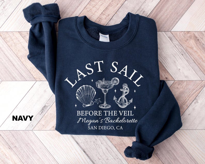 May include: Navy blue sweatshirt with white text and graphics. The text reads "LAST SAIL BEFORE THE VEIL" with a seashell, cocktail, and anchor graphic. The text "Megan's Bachelorette SAN DIEGO, CA" is also included.