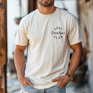May include: Cream-colored t-shirt with the text "COOL Grandpas CLUB" in a circular design. The shirt is a relaxed fit and has short sleeves. The person is wearing a gold watch.