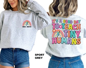 Pode incluir: Uma camisa de moletom cinza com o texto "It's a good day to teach tiny humans" em cores arco-íris nas costas. A frente da camisa de moletom tem um arco-íris com um raio e o texto "Mrs. Smith".