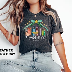 May include: Heather dark gray t-shirt with a colorful illustration of the nativity scene. The text "The greatest gift of all" and "Matthew 1:21" are printed below the illustration.