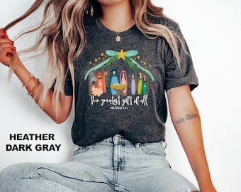 Puede incluir: Camiseta gris oscuro jaspeada con una ilustración colorida de la escena de la natividad. El texto "The greatest gift of all" y "Matthew 1:21" están impresos debajo de la ilustración.