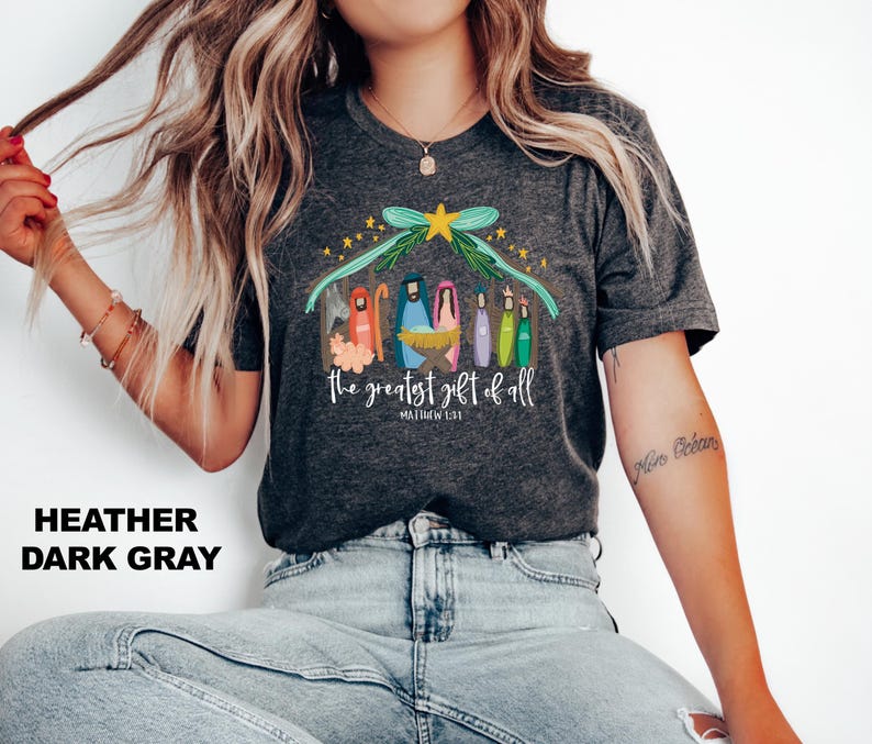 May include: Heather dark gray t-shirt with a colorful illustration of the nativity scene. The text "The greatest gift of all" and "Matthew 1:21" are printed below the illustration.
