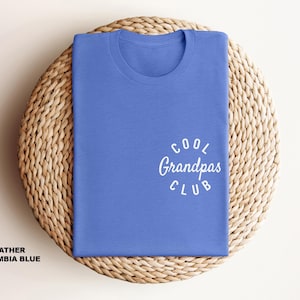 May include: A folded heather Columbia blue t-shirt with the text "Cool Grandpas Club" in white script. The shirt is displayed on a woven, circular placemat. The text "Heather Columbia Blue" is at the bottom.