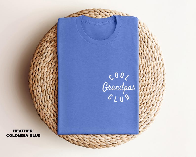 May include: A folded heather Columbia blue t-shirt with the text "Cool Grandpas Club" in white script. The shirt is displayed on a woven, circular placemat. The text "Heather Columbia Blue" is at the bottom.