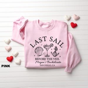 May include: Pink sweatshirt with the text "LAST SAIL BEFORE THE VEIL" and a seashell, cocktail, and anchor graphic. The text "Megan's Bachelorette" and "San Diego, CA" are also printed on the shirt.