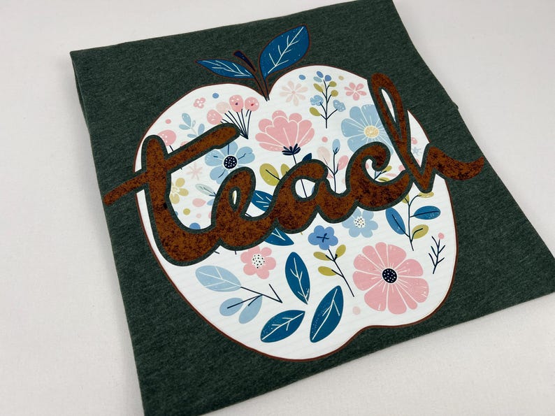 Op de afbeelding: Een donkergroen T-shirt met een wit appelontwerp met een bloemenpatroon en het woord "teach" in bruine letters.