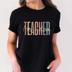 K&ouml;nnte beinhalten: Schwarzes T-Shirt mit dem Text "TEACHER" in bunten Buchstaben und "Mrs. Wilson" in wei&szlig;er Schrift darunter.