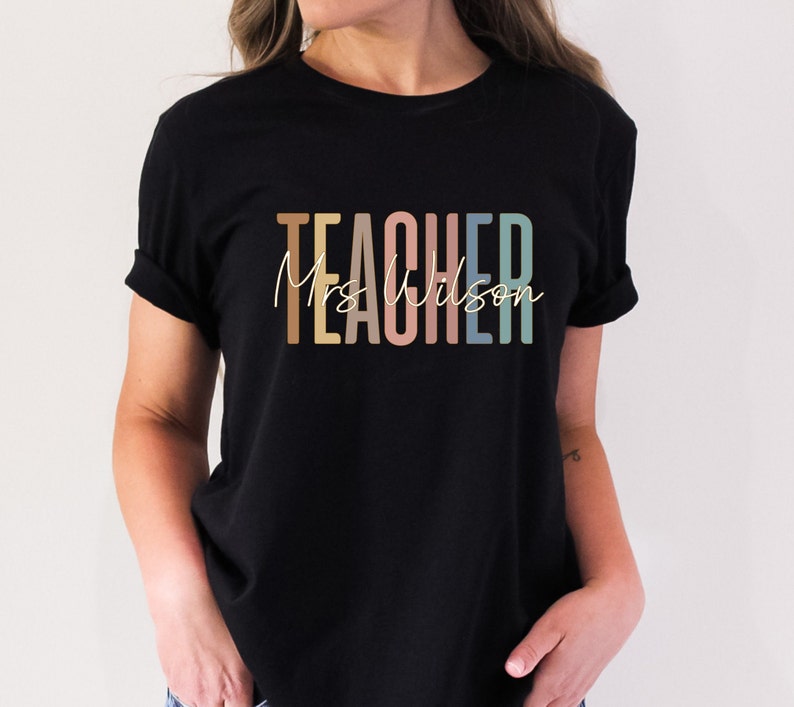K&ouml;nnte beinhalten: Schwarzes T-Shirt mit dem Text "TEACHER" in bunten Buchstaben und "Mrs. Wilson" in wei&szlig;er Schrift darunter.