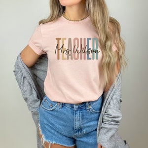 K&ouml;nnte beinhalten: Ein rosa T-Shirt mit dem Wort "TEACHER" in Regenbogenfarben und dem Text "Mrs. Wilson" in schwarzer Schrift.