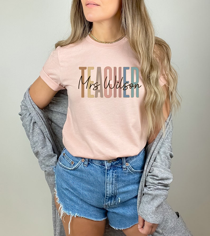 K&ouml;nnte beinhalten: Ein rosa T-Shirt mit dem Wort "TEACHER" in Regenbogenfarben und dem Text "Mrs. Wilson" in schwarzer Schrift.
