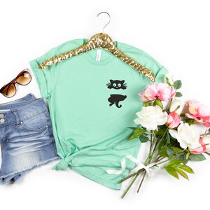 May include: A mint green t-shirt with a black cat graphic on the front pocket. The shirt is on a gold hanger and is surrounded by a pair of sunglasses, denim shorts, and a bouquet of white and pink flowers.