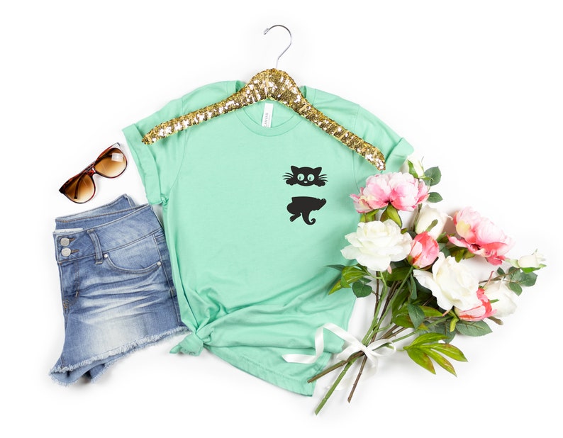 May include: A mint green t-shirt with a black cat graphic on the front pocket. The shirt is on a gold hanger and is surrounded by a pair of sunglasses, denim shorts, and a bouquet of white and pink flowers.