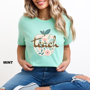 Op de afbeelding: Een mintgroen T-shirt met een bloemachtig appelontwerp en het woord "teach" in bruine letters.
