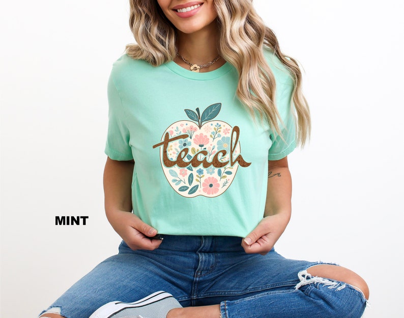Op de afbeelding: Een mintgroen T-shirt met een bloemachtig appelontwerp en het woord "teach" in bruine letters.