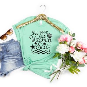 May include: A mint green t-shirt with the text "All I need is Vitamin Sea" in black. The text is surrounded by a seashell, a starfish, and wavy lines. The t-shirt is on a white background with a pair of sunglasses, denim shorts, and a bouquet of pink and white flowers.