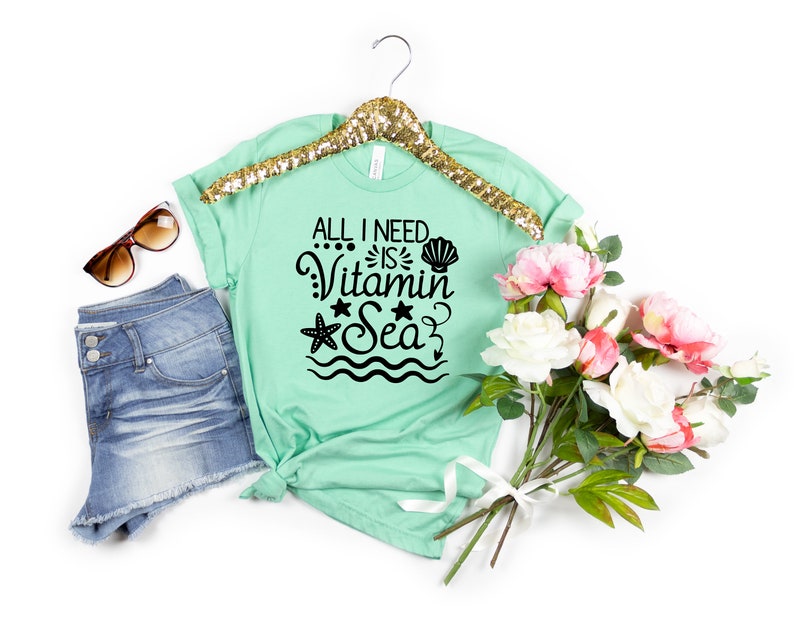 May include: A mint green t-shirt with the text "All I need is Vitamin Sea" in black. The text is surrounded by a seashell, a starfish, and wavy lines. The t-shirt is on a white background with a pair of sunglasses, denim shorts, and a bouquet of pink and white flowers.