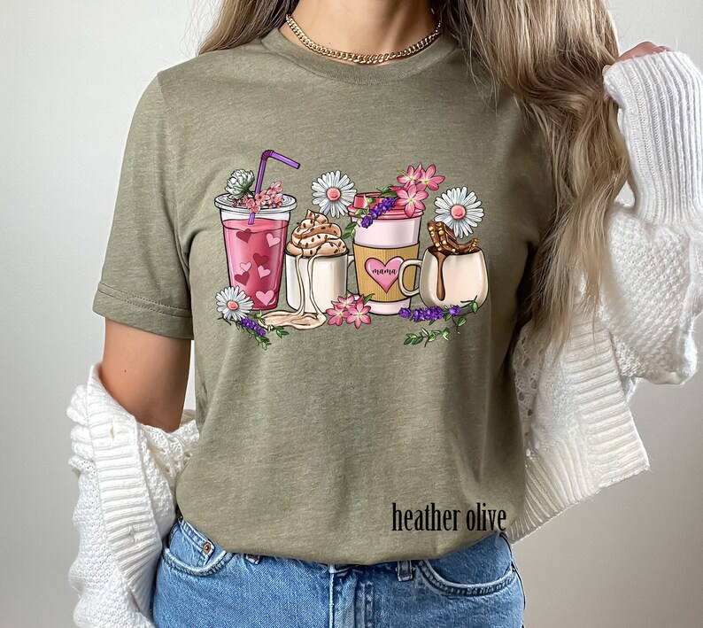 Pode incluir: Uma camiseta verde com um gr&aacute;fico de quatro bebidas de caf&eacute; diferentes com flores e a palavra "Happiness" em uma das x&iacute;caras. O texto "heather olive" est&aacute; impresso na parte inferior da camiseta.