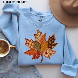 Pode incluir: Uma camisola de gola redonda azul-clara com uma grande aplica&ccedil;&atilde;o de folha de &aacute;cer ao centro, feita de v&aacute;rios retalhos de tecido em tons de outono como laranja, castanho, verde e amarelo, com costuras decorativas vis&iacute;veis nas margens.