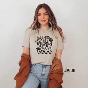 May include: A tan t-shirt with the text "All I need is Vitamin Sea" in black. The text is surrounded by a seashell, a starfish, and wavy lines.