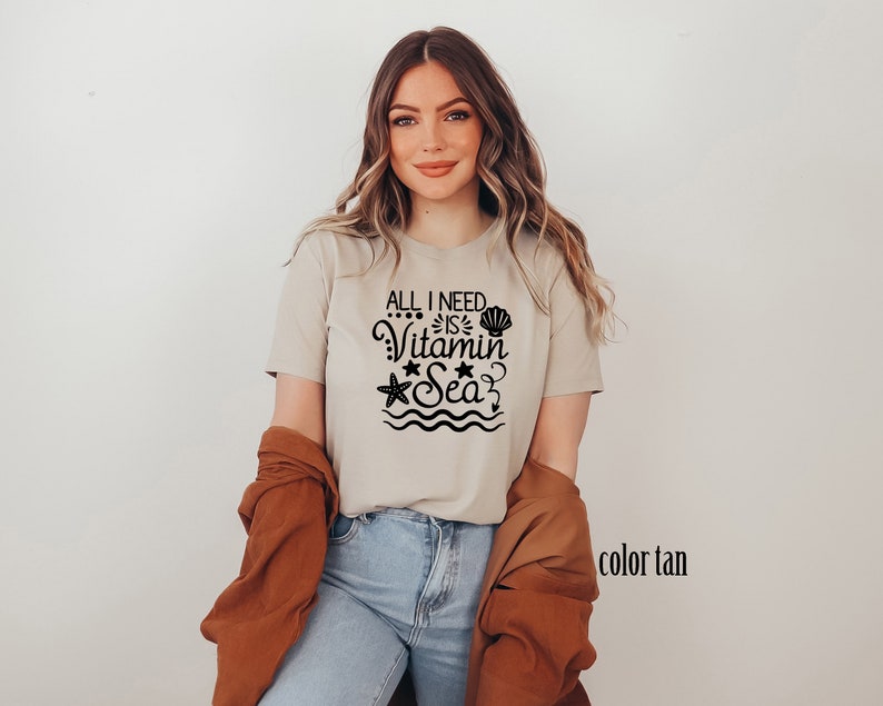 May include: A tan t-shirt with the text "All I need is Vitamin Sea" in black. The text is surrounded by a seashell, a starfish, and wavy lines.