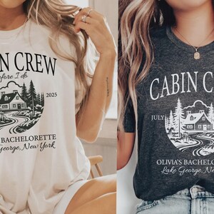 May include: A cream t-shirt and a dark grey t-shirt, each featuring a graphic of a cabin in the woods with the text "CABIN CREW before I do", "JULY 2025", "OLIVIA'S BACHELORETTE", and "Lake George, New York" printed in a circular design.