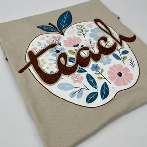 Op de afbeelding: Een beige t-shirt met een bloemachtig appelontwerp en het woord "teach" in bruine letters.
