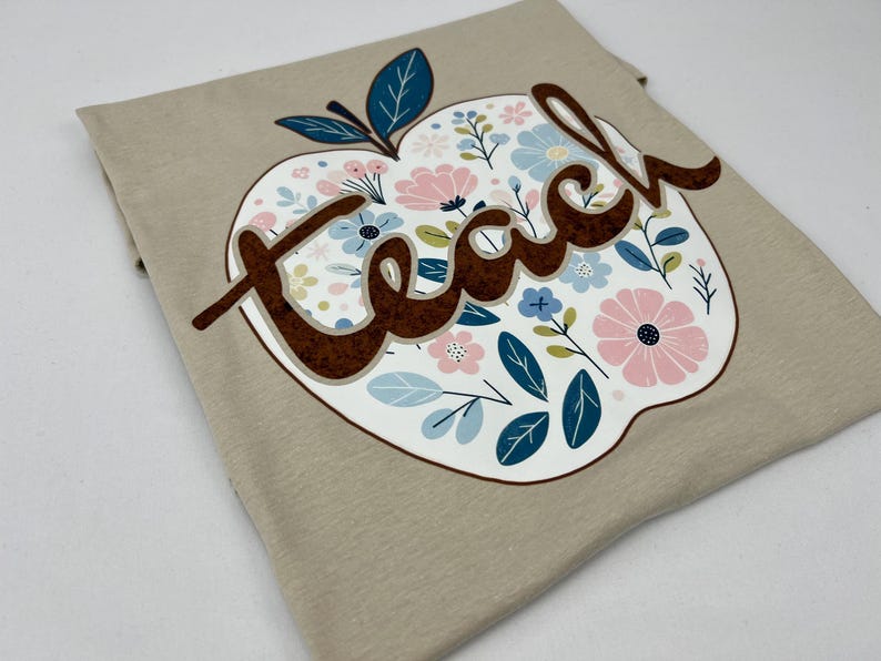 Op de afbeelding: Een beige t-shirt met een bloemachtig appelontwerp en het woord "teach" in bruine letters.