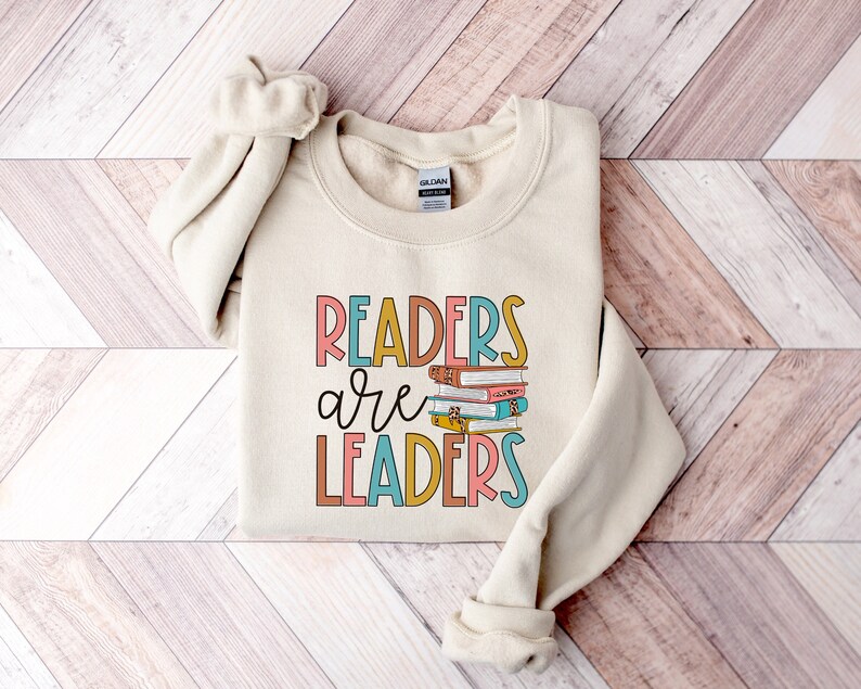 May include: A beige sweatshirt with the text "Readers are Leaders" in colorful letters. A stack of colorful books is pictured above the word "Leaders".