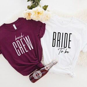 May include: Two t-shirts, one maroon and one white, with text printed on them. The maroon shirt says "back crew" in white text. The white shirt says "bride to be" in black text. A bottle of sparkling wine is in the center of the image.