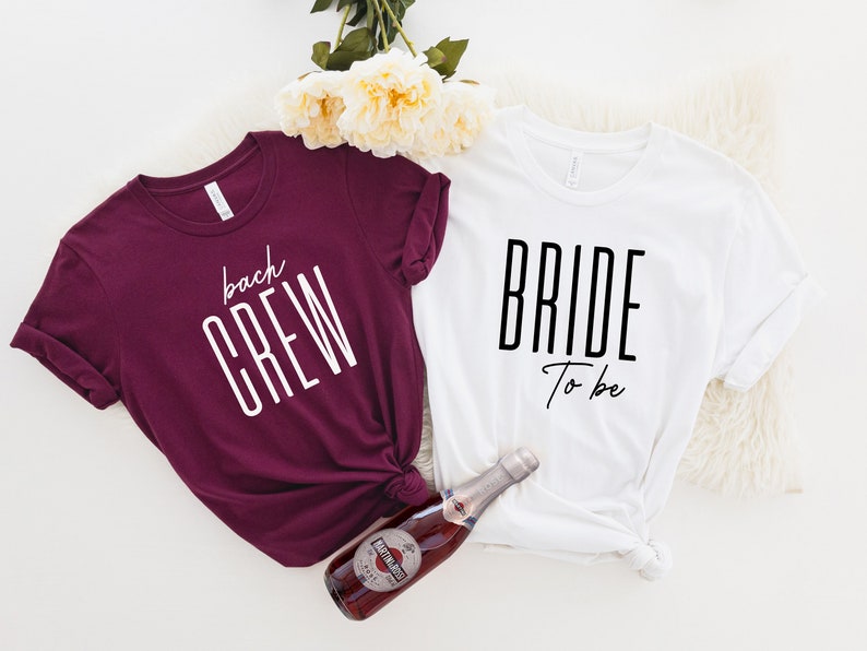 May include: Two t-shirts, one maroon and one white, with text printed on them. The maroon shirt says "back crew" in white text. The white shirt says "bride to be" in black text. A bottle of sparkling wine is in the center of the image.
