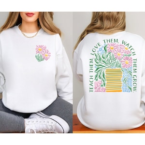 May include: White sweatshirt with a floral design. The front features a small cluster of pink daisies with green leaves. The back displays a larger floral arrangement in a vase, with the text "Teach them, love them, watch them grow."