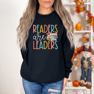 May include: Black sweatshirt with the text "Readers are Leaders" in colorful letters. A stack of colorful books is pictured next to the text.