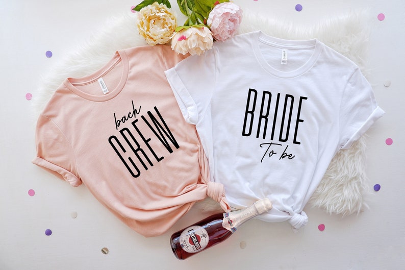 May include: Two white t-shirts with black text. One shirt says "Bride to be" and the other says "bach crew".