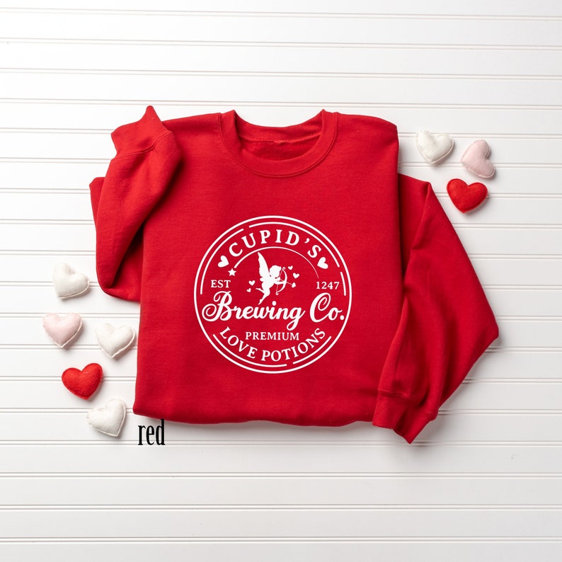 May include: Red sweatshirt with a white graphic of a vintage logo that says "Cupid's Brewing Co. Est. 1247 Premium Love Potions". The logo features a cupid with an arrow.