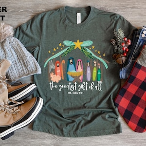 May include: A green t-shirt with a Christmas nativity scene design. The design features a manger with a star above it and the words "The greatest gift of all" with the scripture reference "Matthew 1:21".