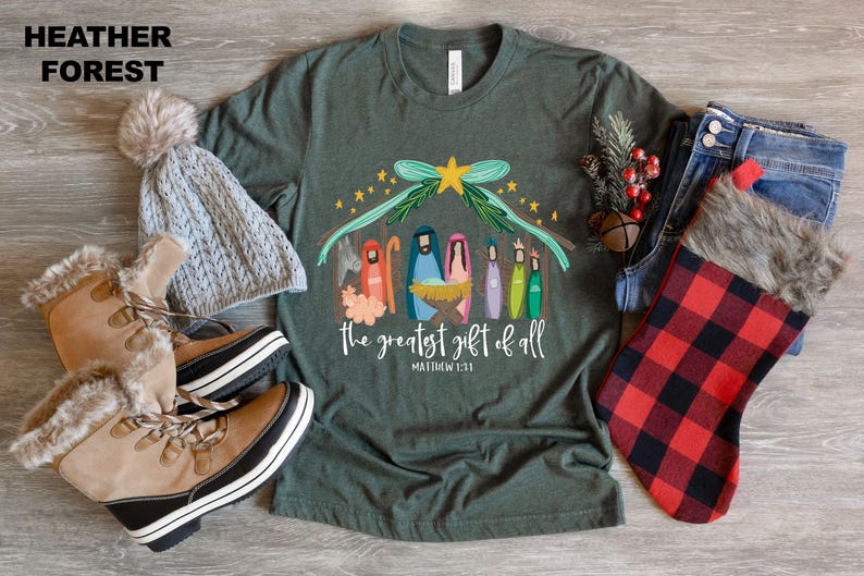 May include: A green t-shirt with a Christmas nativity scene design. The design features a manger with a star above it and the words "The greatest gift of all" with the scripture reference "Matthew 1:21".