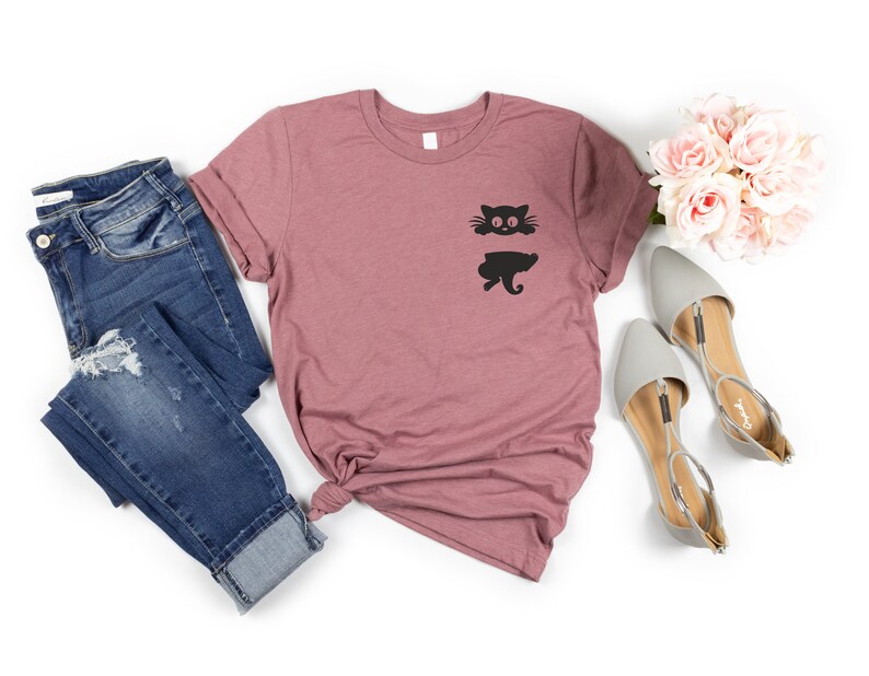 May include: A mauve t-shirt with a black cat graphic on the front. The cat is split in half, with the top half showing the head and the bottom half showing the tail and legs. The t-shirt is paired with blue jeans and gray sandals.