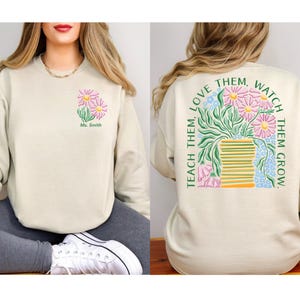 May include: Beige sweatshirt with a floral design. The front features a small bouquet of pink and yellow flowers with the text "Ms. Smith". The back displays a larger floral arrangement with the text "Teach them, love them, watch them grow."